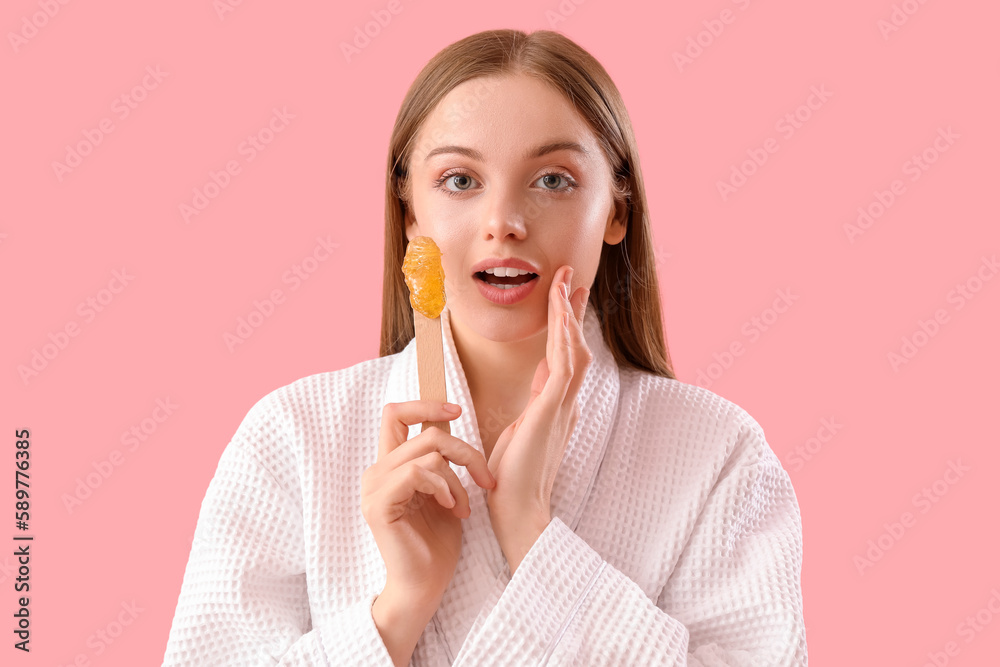 Surprised young woman holding spatula with sugaring paste on pink background