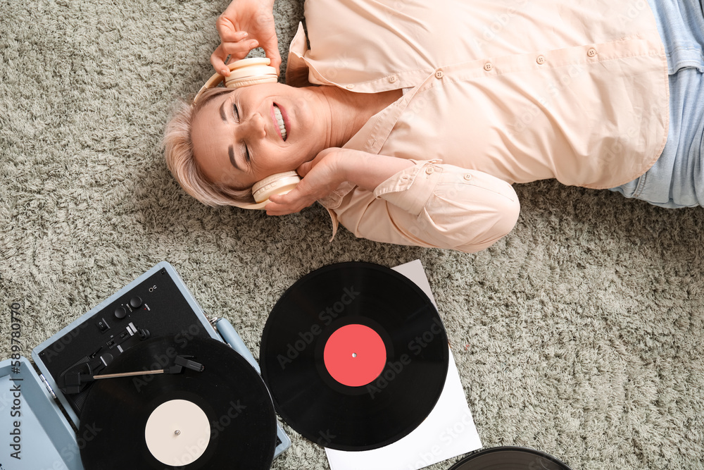 Mature woman in headphones with record player lying on carpet at home, top view