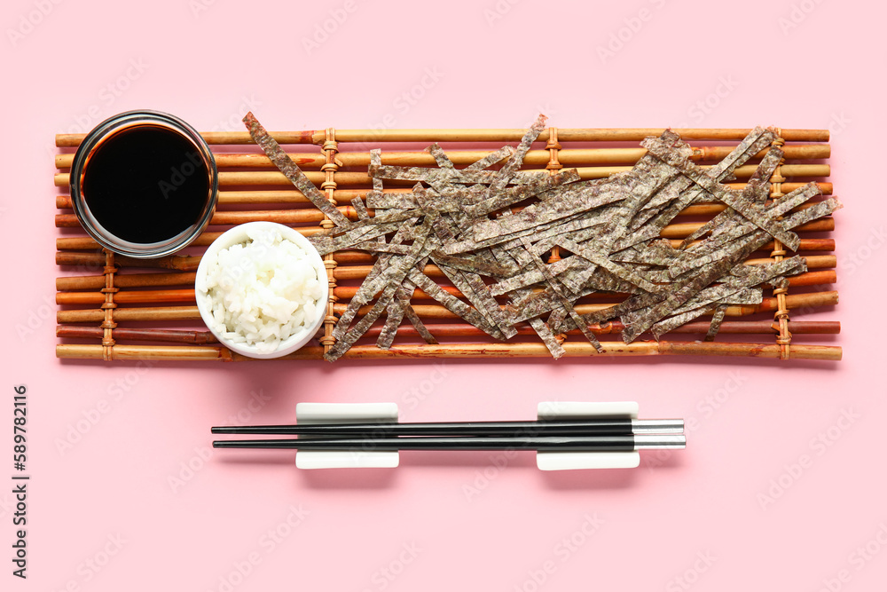 Bamboo mat with cut nori sheets, rice, soy sauce and chopsticks on pink background