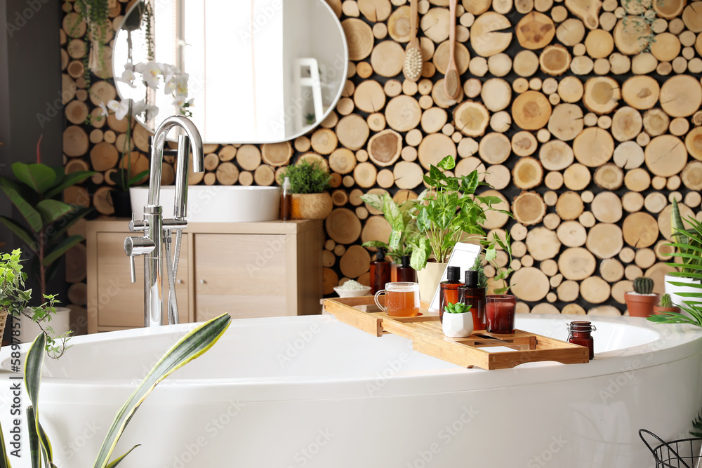 Stand with cosmetic products, cup, tablet computer and houseplants on bathtub in room