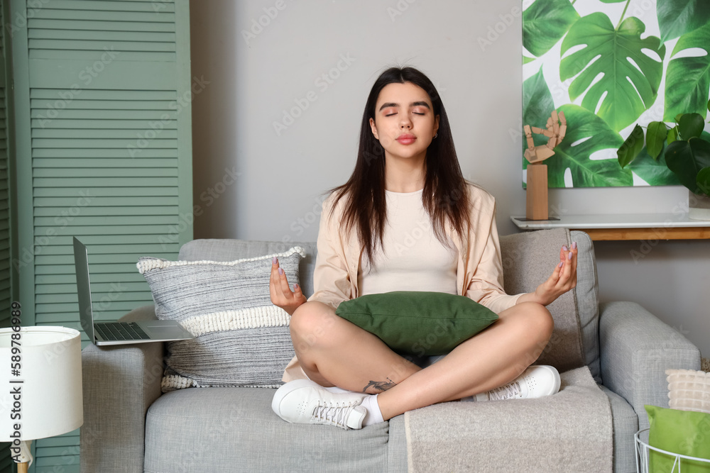 Young woman meditating with laptop on sofa at home