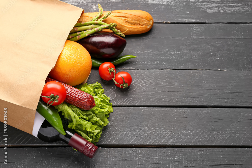 Paper bag with vegetables, sausage and wine bottle on black wooden background