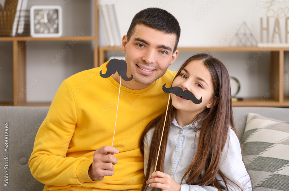 Portrait of father and his little daughter with paper mustache sitting on sofa