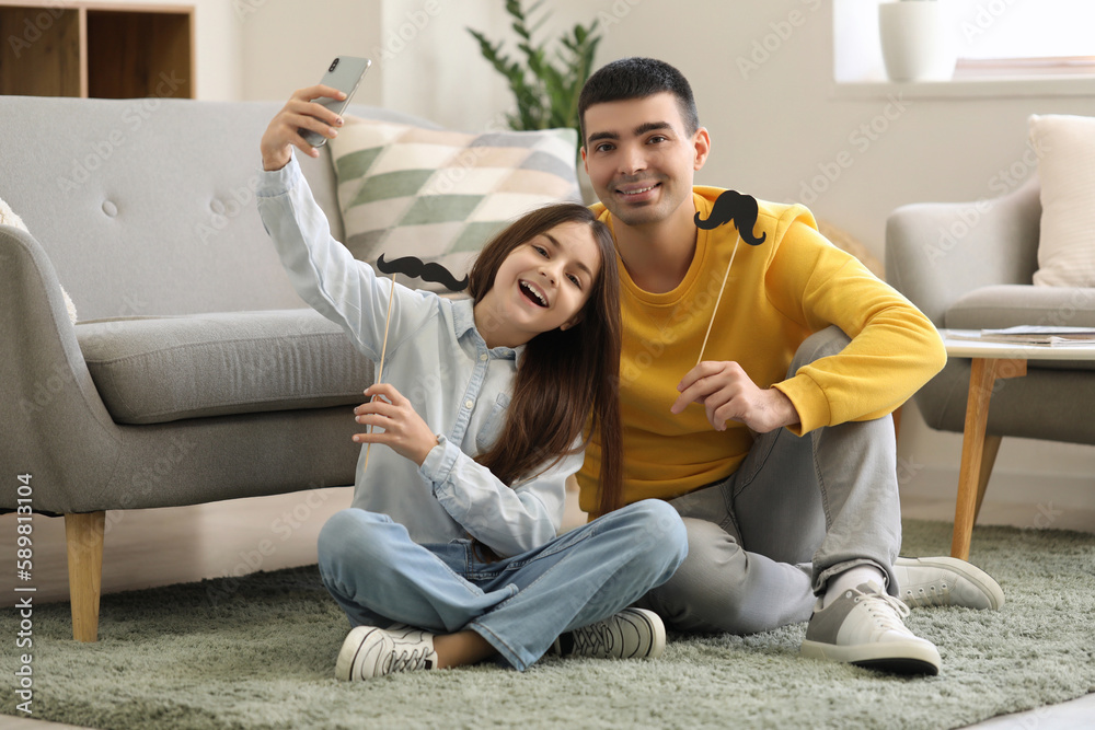 Portrait of father and his little daughter with paper mustache taking selfie at home