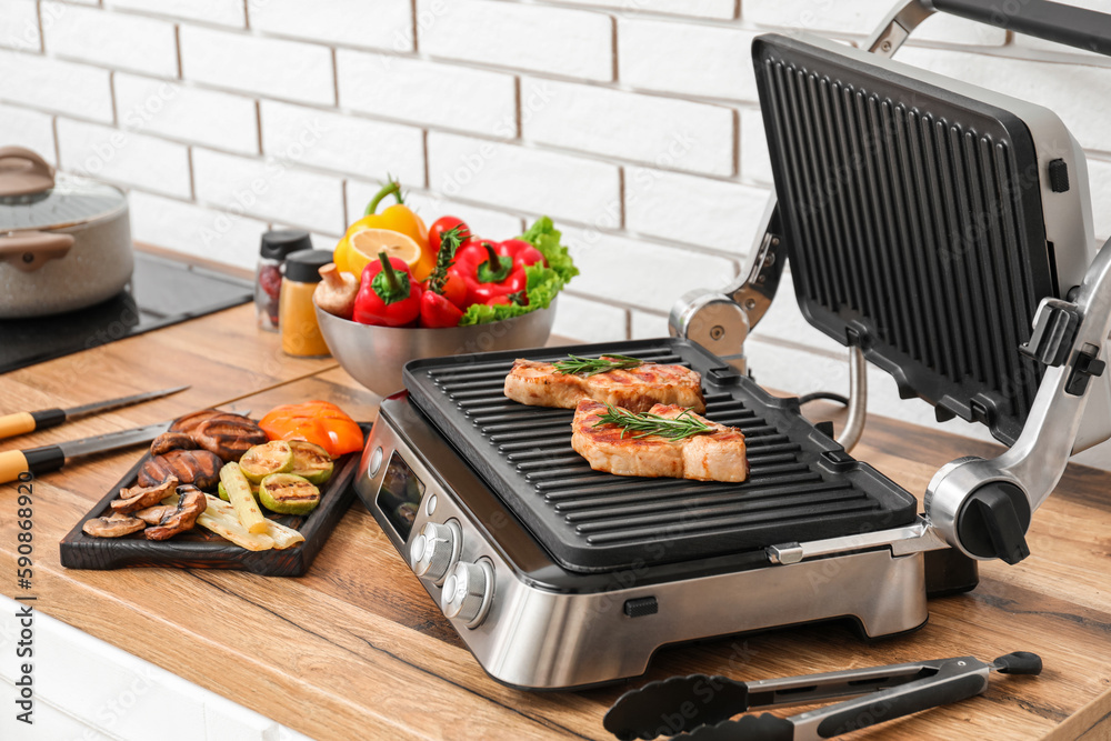 Modern electric grill with tasty steaks and vegetables on table near light brick wall