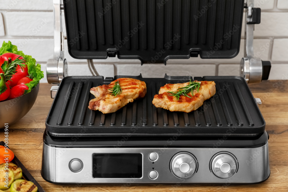 Modern electric grill with tasty steaks on table near light brick wall