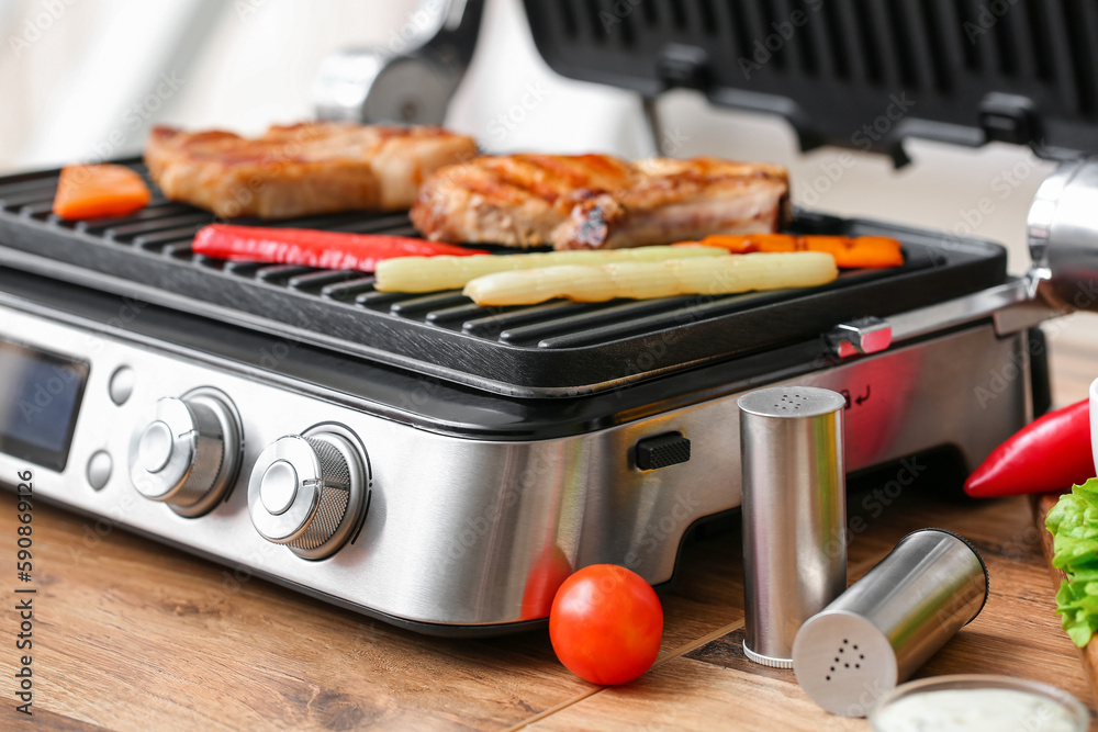 Modern electric grill with tasty steaks and vegetables on table, closeup