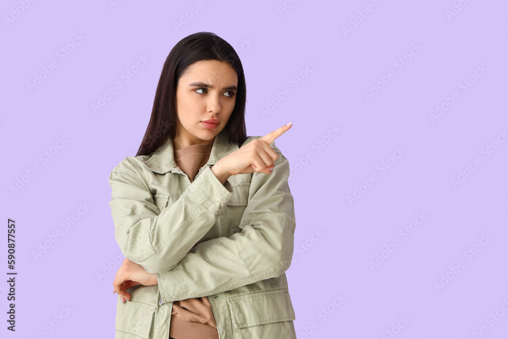 Displeased young woman pointing at something on lilac background