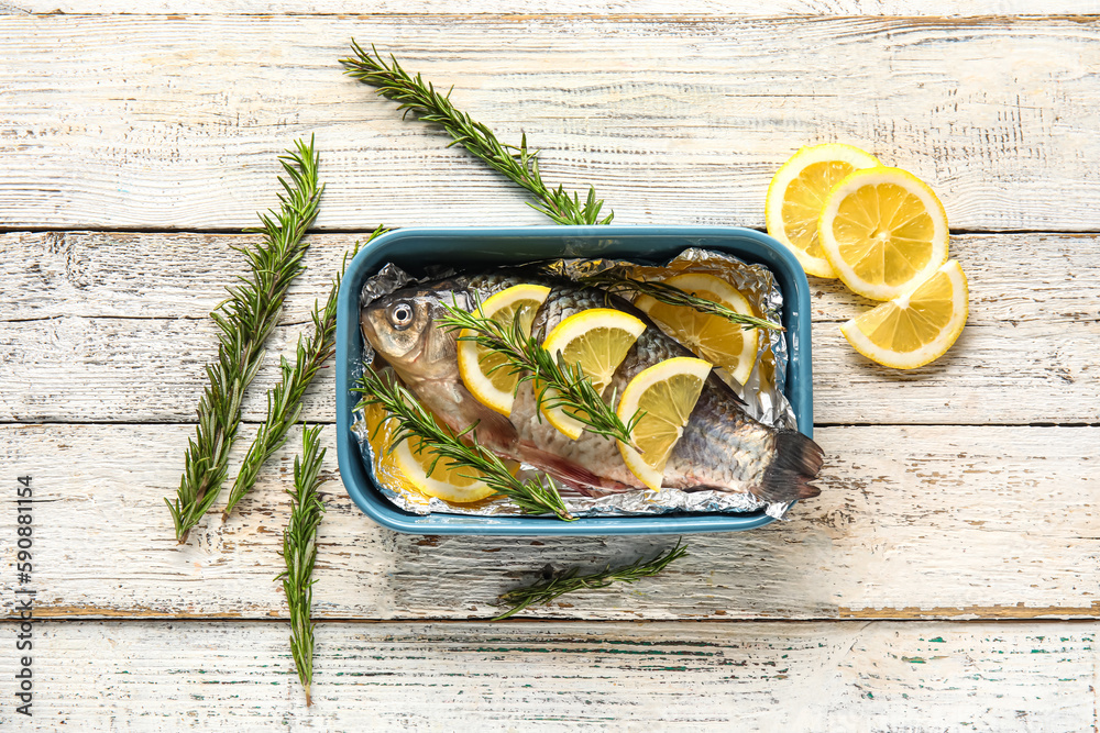 Baking dish with raw fish, lemon slices and rosemary on light wooden background