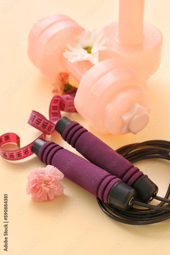 Dumbbells with skipping rope, tape measure and spring flowers on yellow background, closeup