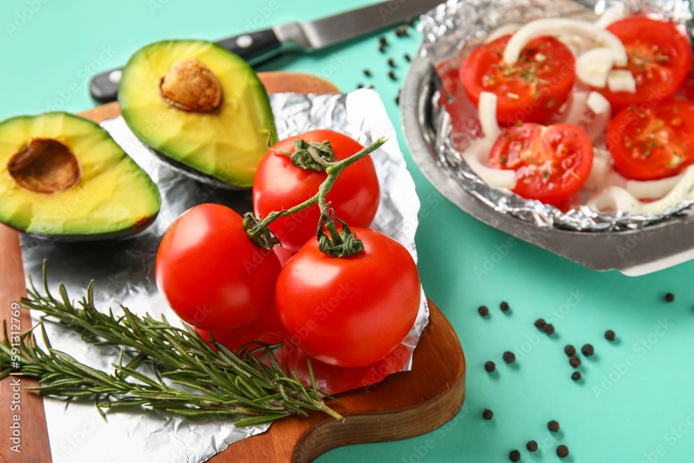 Wooden board with aluminium foil, fresh tomatoes, avocado and rosemary on color background, closeup