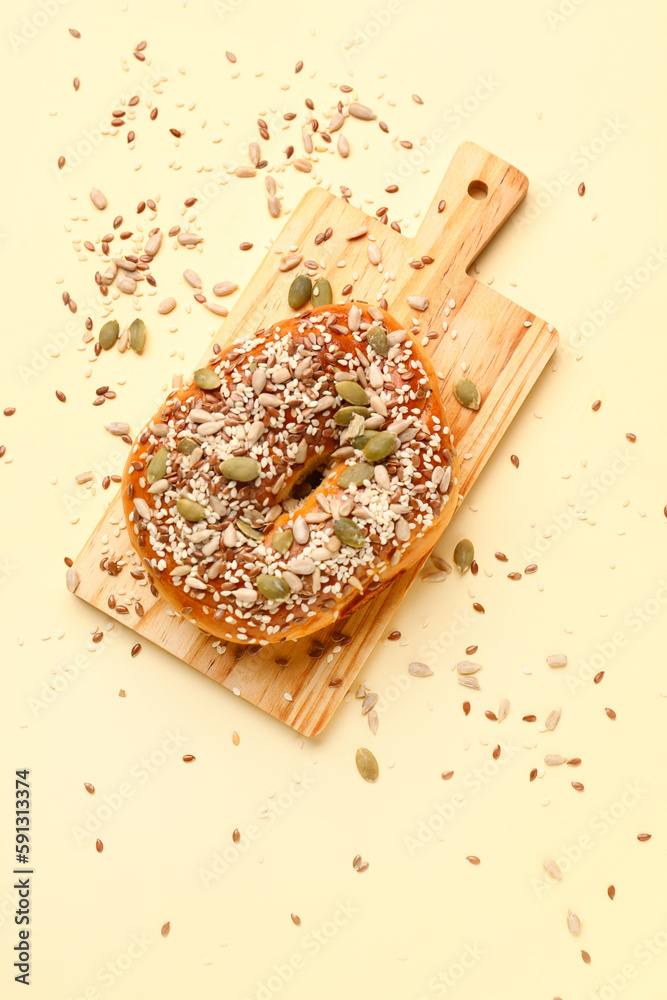 Wooden board of tasty bagel with sesame and pumpkin seeds on beige background