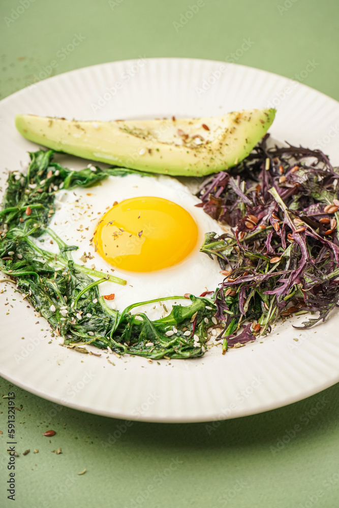 Plate with tasty fried egg, avocado and greens on green background