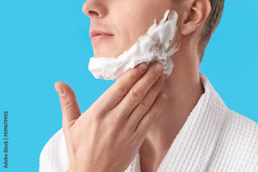 Young man applying shaving foam onto face against light blue background, closeup