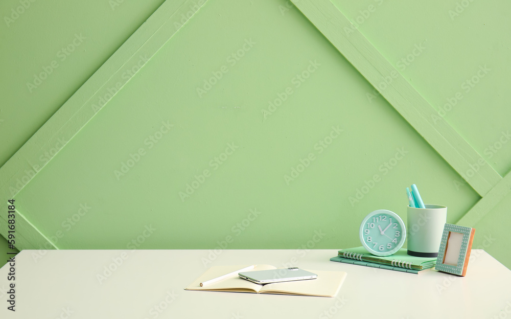 Workplace with stationery, mobile phone and alarm clock near green wall