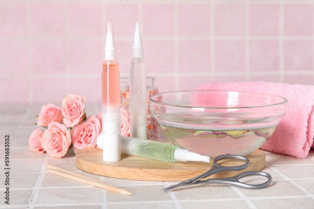 Wooden board with cuticle oil pens, manicure instruments, bowl of water and rose flowers on light ti