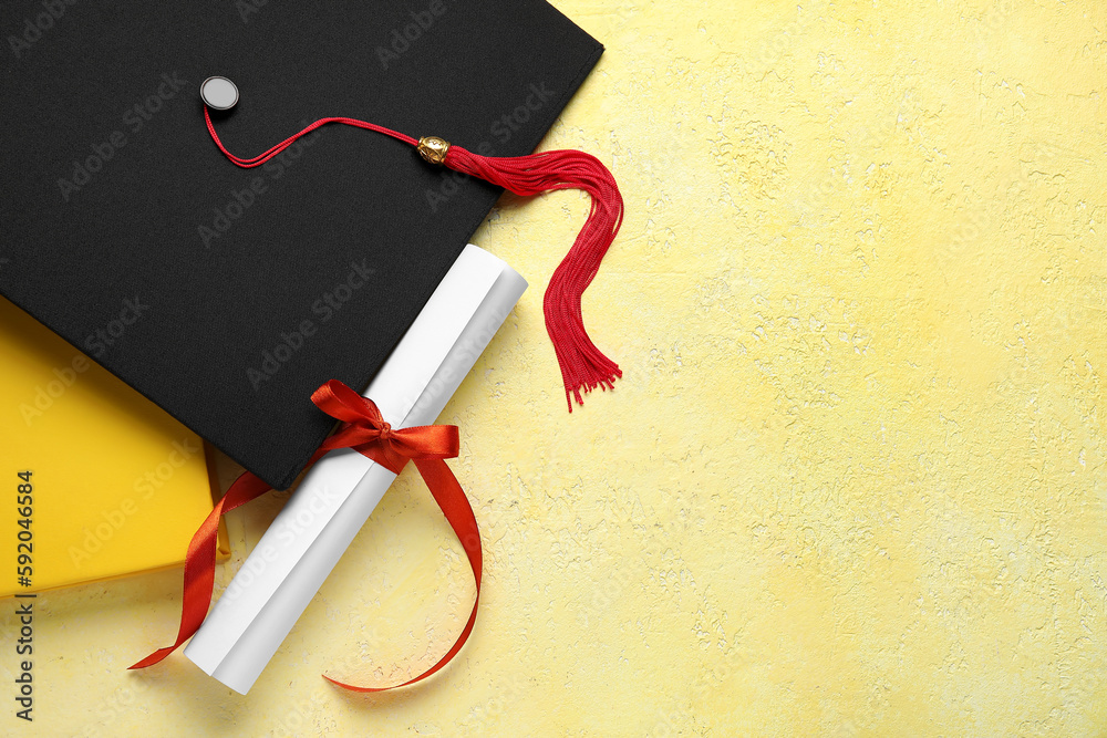 Diploma with red ribbon, graduation hat and book on yellow table