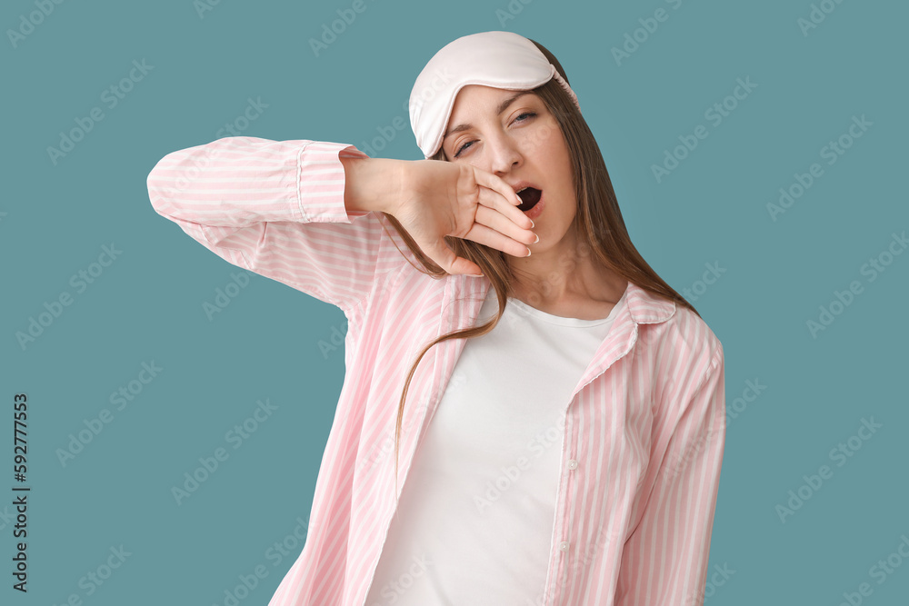 Young woman with sleeping mask yawning on blue background