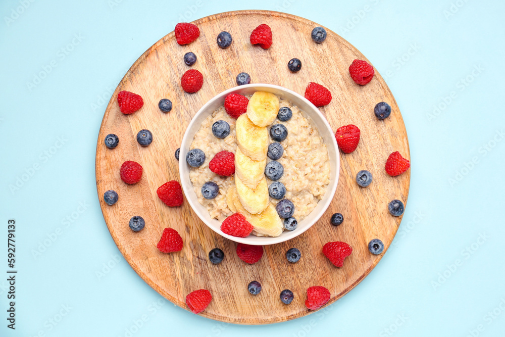 Board with raspberries, blueberries and bowl of tasty oatmeal on blue background