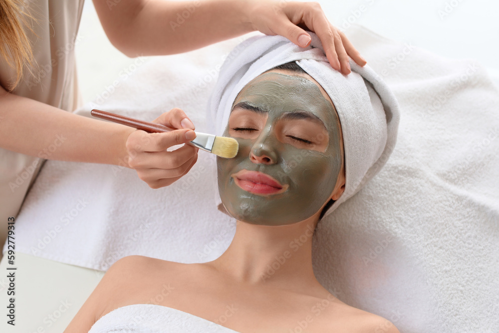 Cosmetologist applying clay mask onto young womans face in salon, closeup