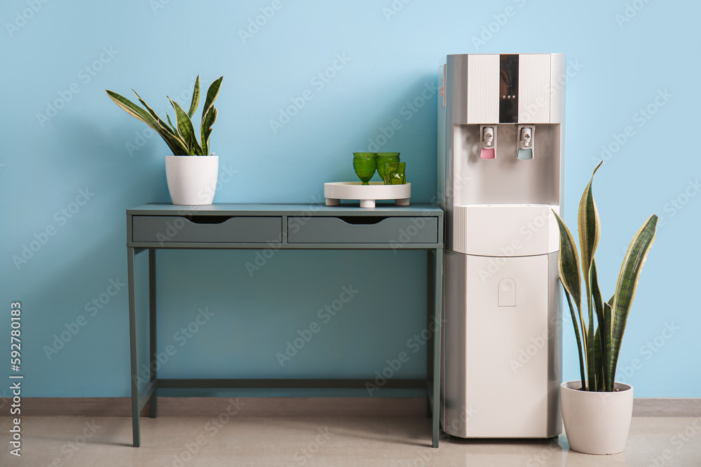 Modern water cooler, table, glasses and houseplants near blue wall