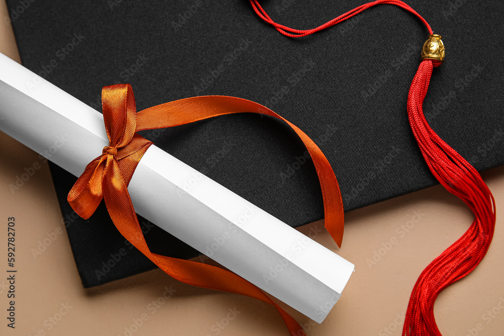 Diploma with red ribbon and graduation hat on beige table