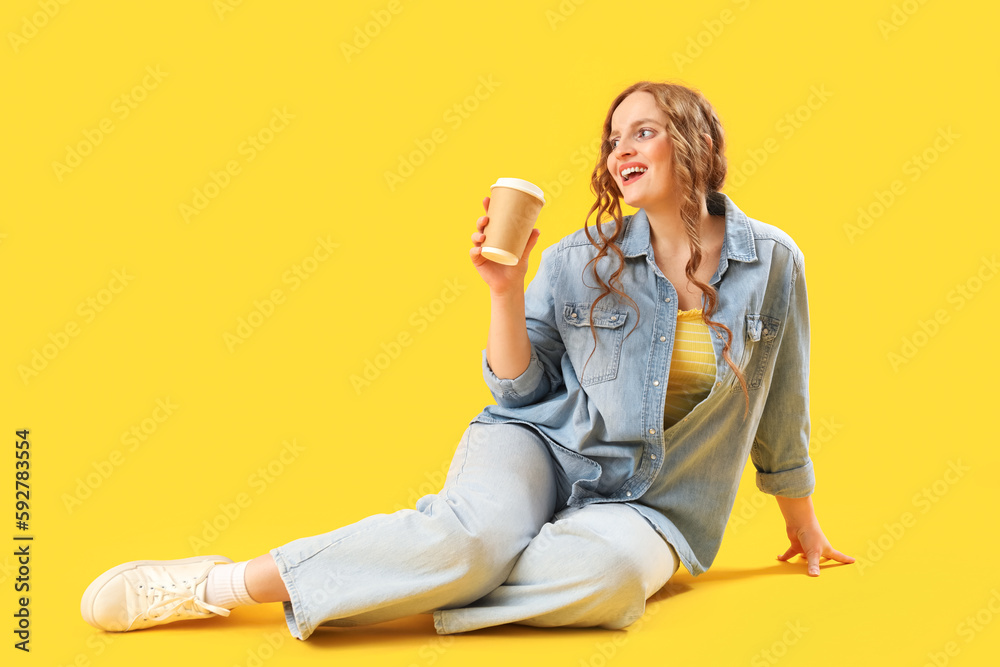 Beautiful redhead woman with cup of coffee sitting on yellow background