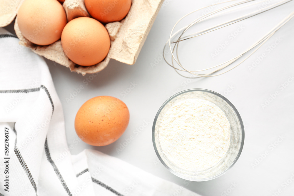 Glass with wheat flour, eggs in package and whisk on white background