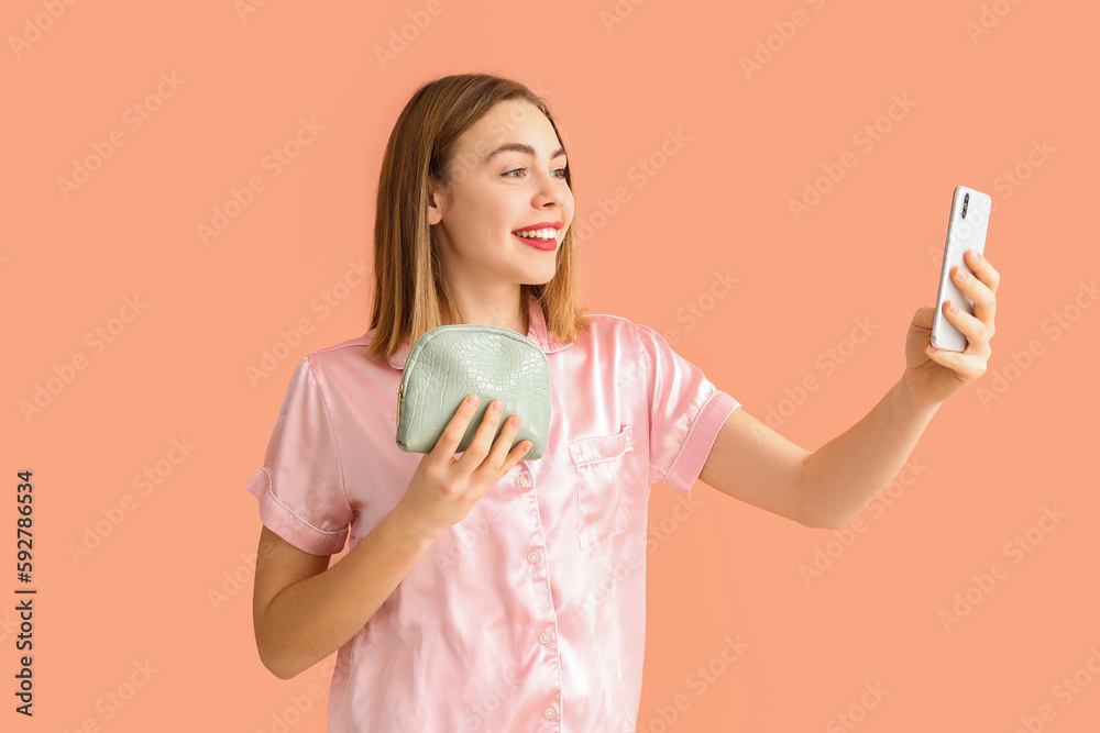 Young woman with cosmetic bag and mobile phone taking selfie on pink background