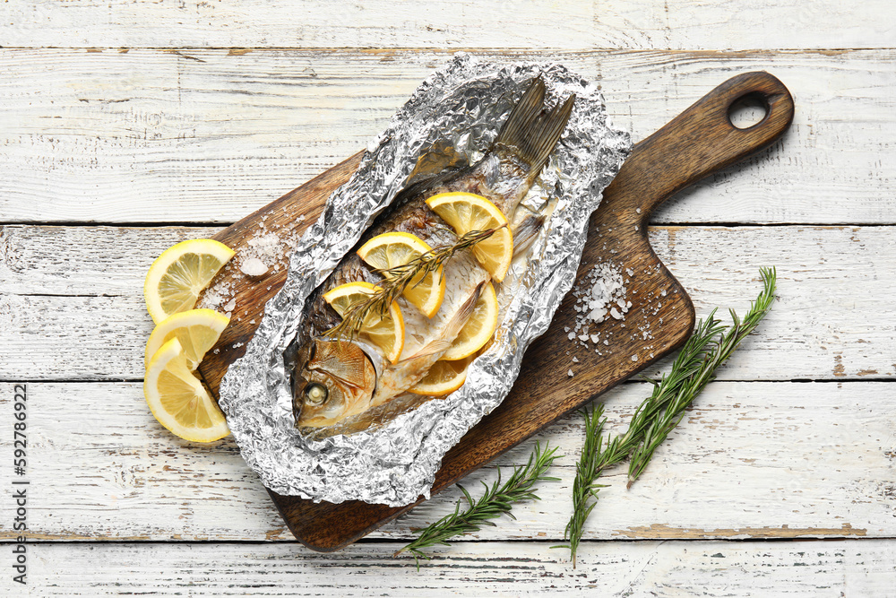 Aluminium foil with tasty baked fish, lemon slices and rosemary on light wooden background