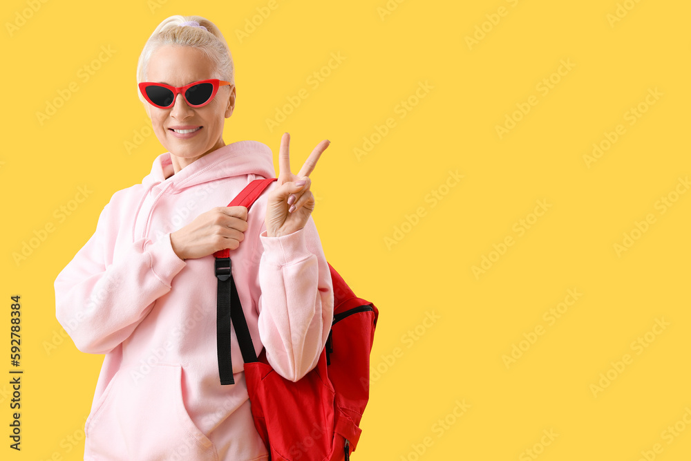 Mature woman with backpack showing victory gesture on yellow background