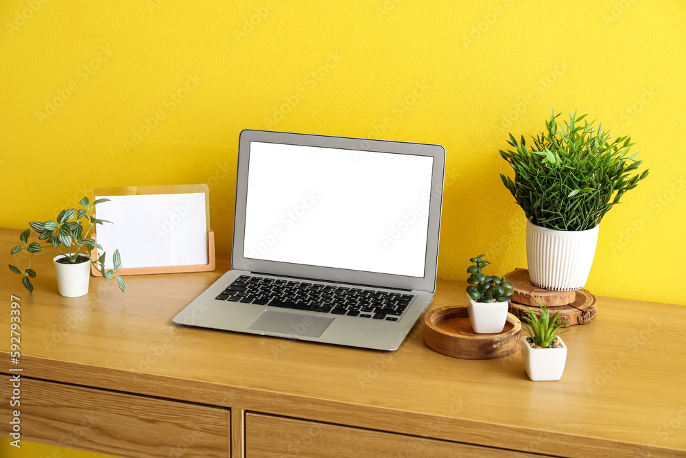 Workplace with artificial plants and laptop near yellow wall