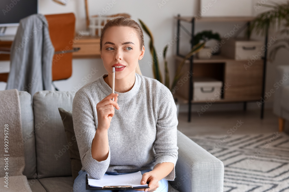 Thoughtful young woman with clipboard sitting on sofa at home