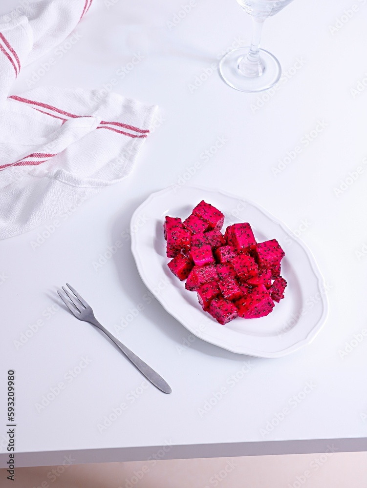 Vertical shot of pink dragon fruit cubes in a white plate with a fork and towel on a table