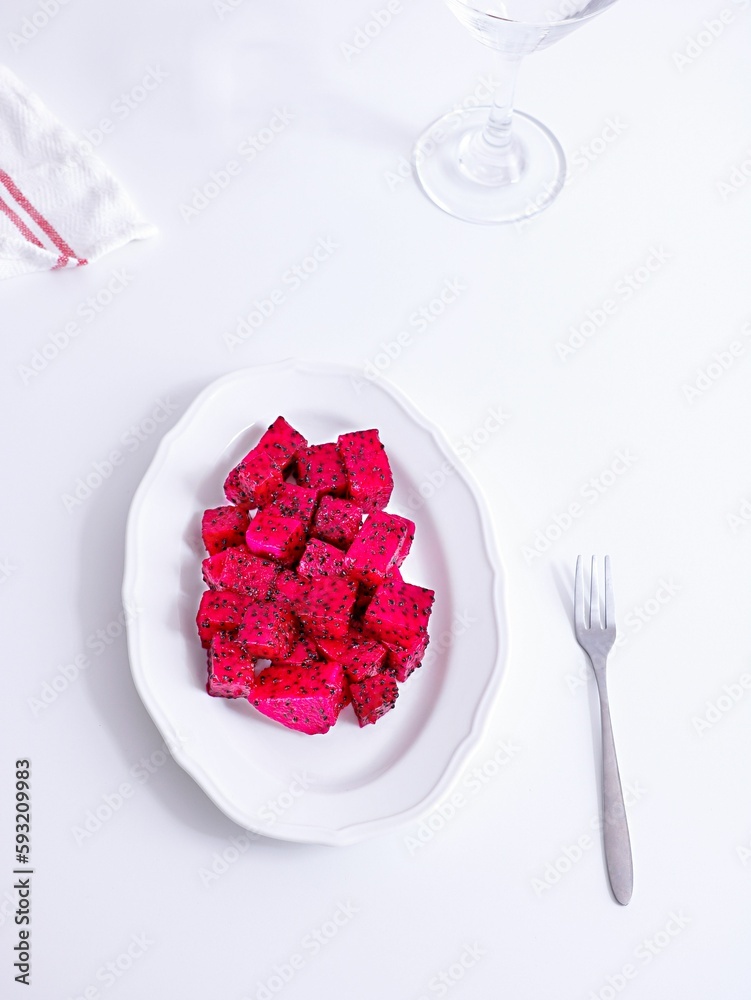 Vertical shot of pink dragon fruit cubes in a white plate with a fork and towel on a table