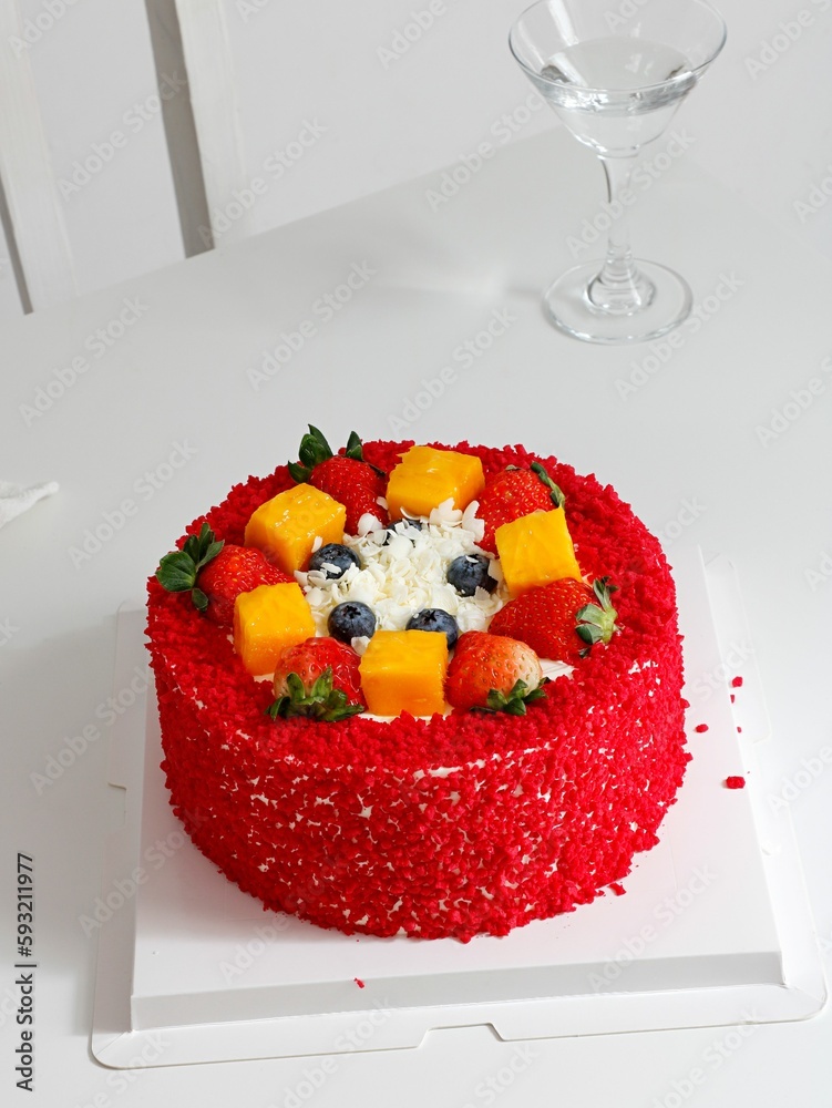 Red, round cake decorated with blueberries, strawberries and mango with a glass of water in the back