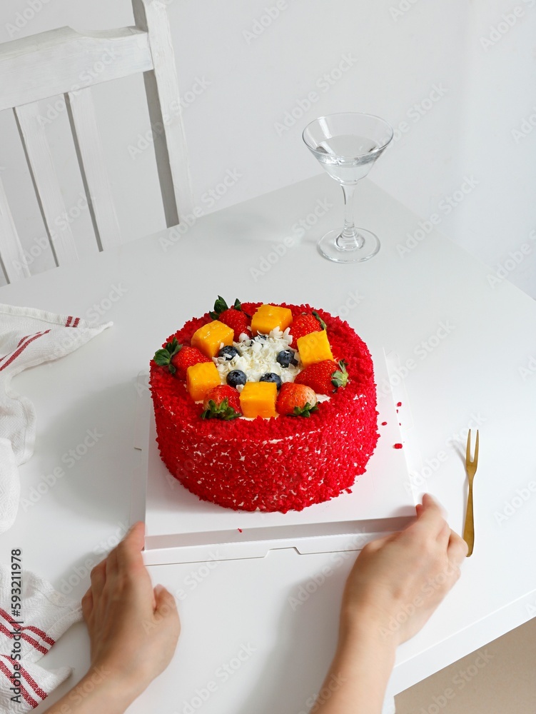 Person holding a red cake with blueberries, strawberries and mango with a glass of water in the back