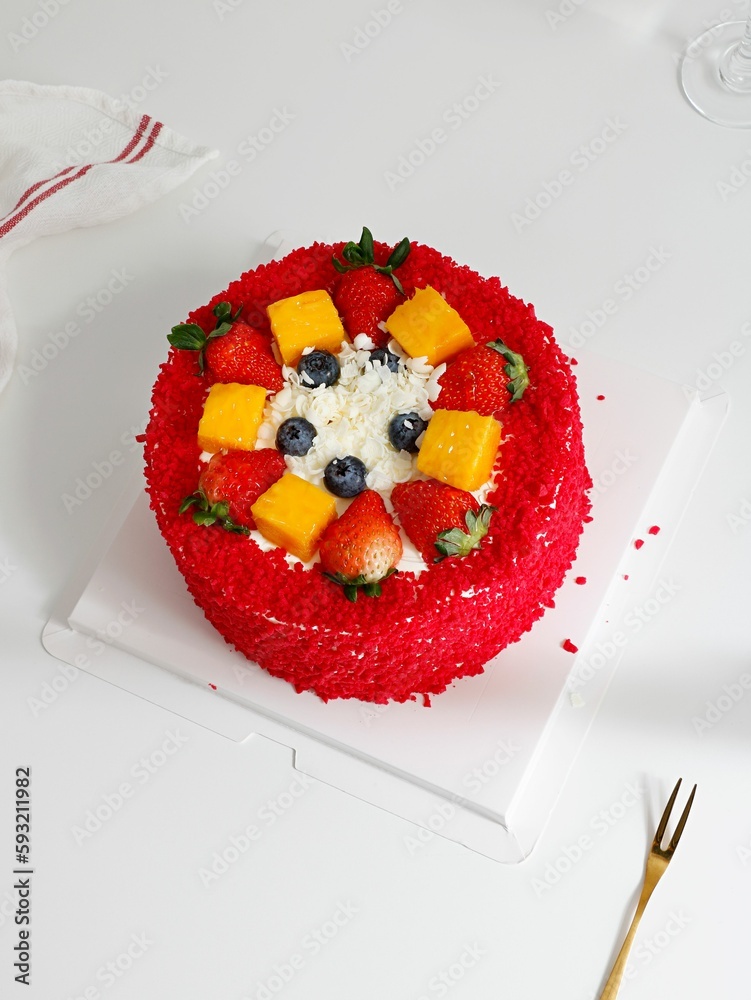 Red, round cake decorated with blueberries, strawberries and mango with a glass of water in the back