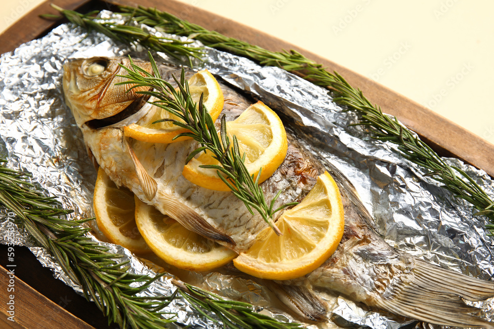 Aluminium foil with tasty baked fish, lemon slices and rosemary on wooden board, closeup