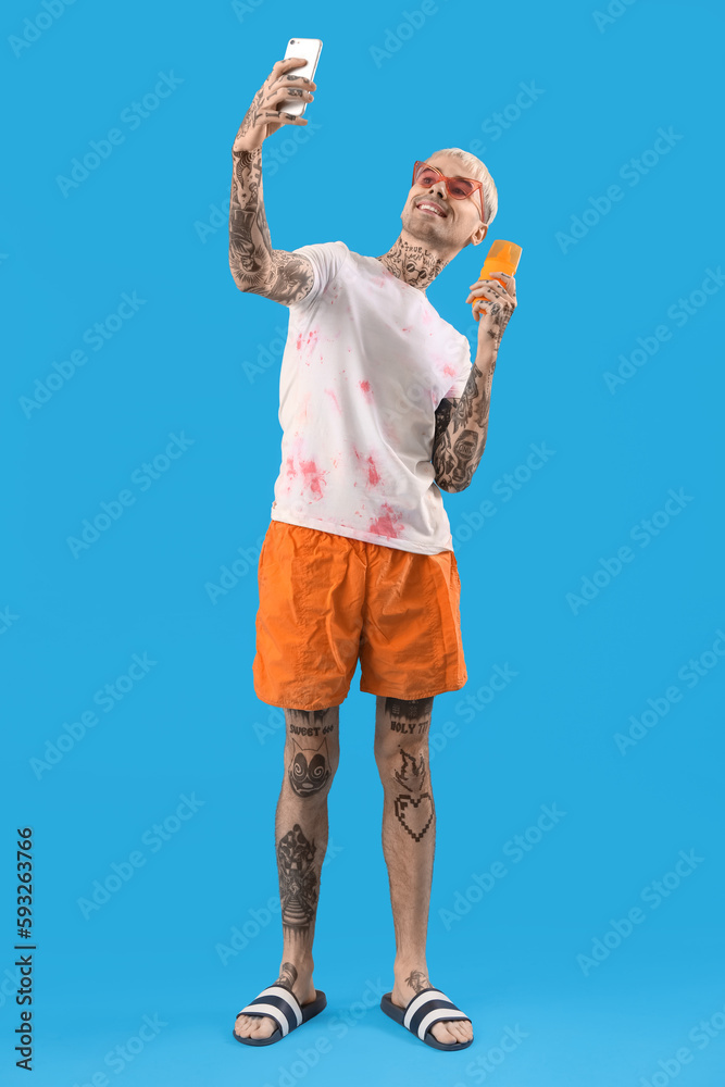 Tattooed man with sunscreen cream taking selfie on light blue background