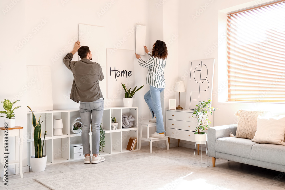 Young couple hanging paintings on light wall at home