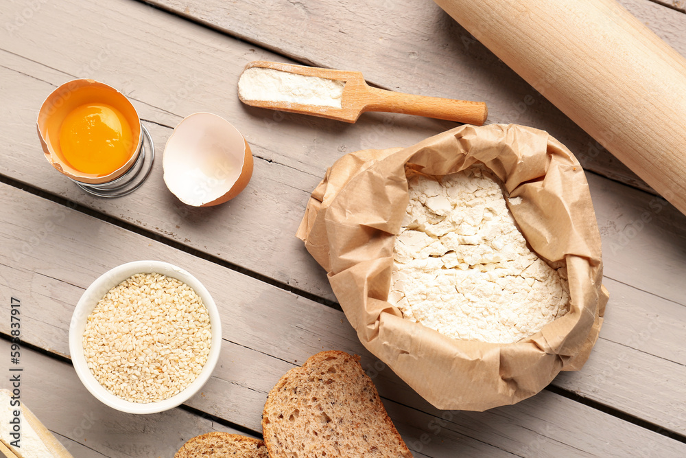 Paper bag of wheat flour, cracked egg, sesame seeds and scoop on grey wooden table