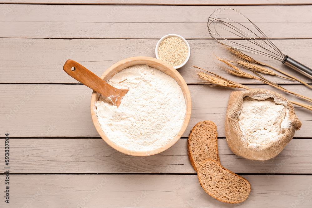 Bowl with wheat flour, bread, sesame seeds and whisk on grey wooden table