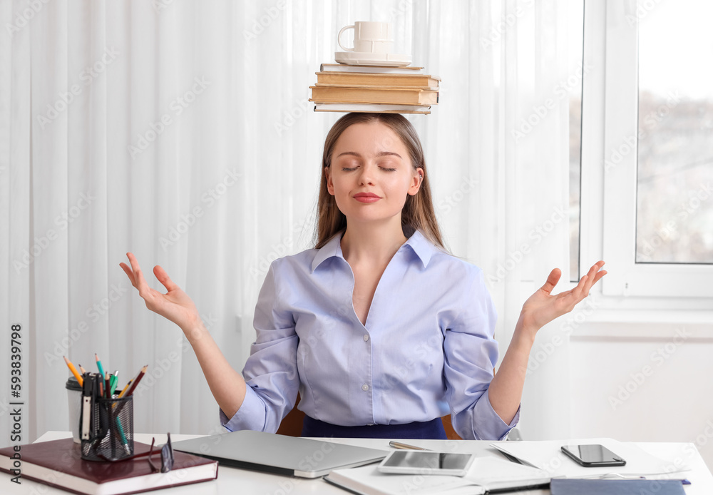 Young businesswoman with cup and books on her head meditating in office. Balance concept