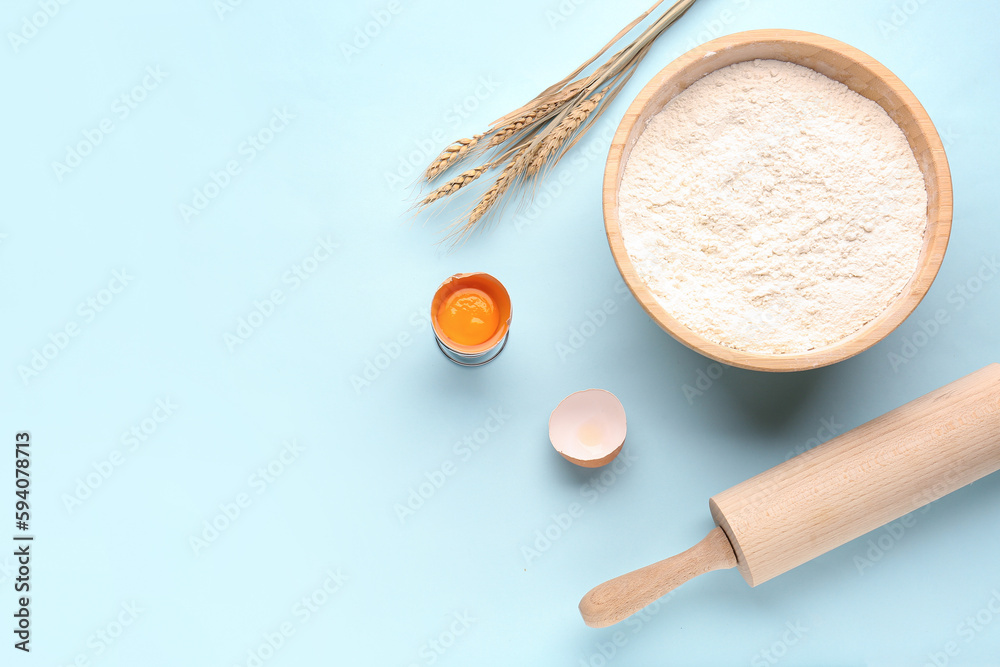 Bowl with flour, wheat ears, cracked egg and rolling pin on blue background