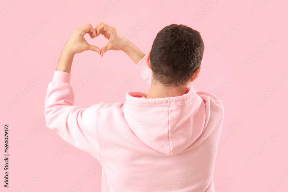 Young man making heart with his hands on pink background, back view