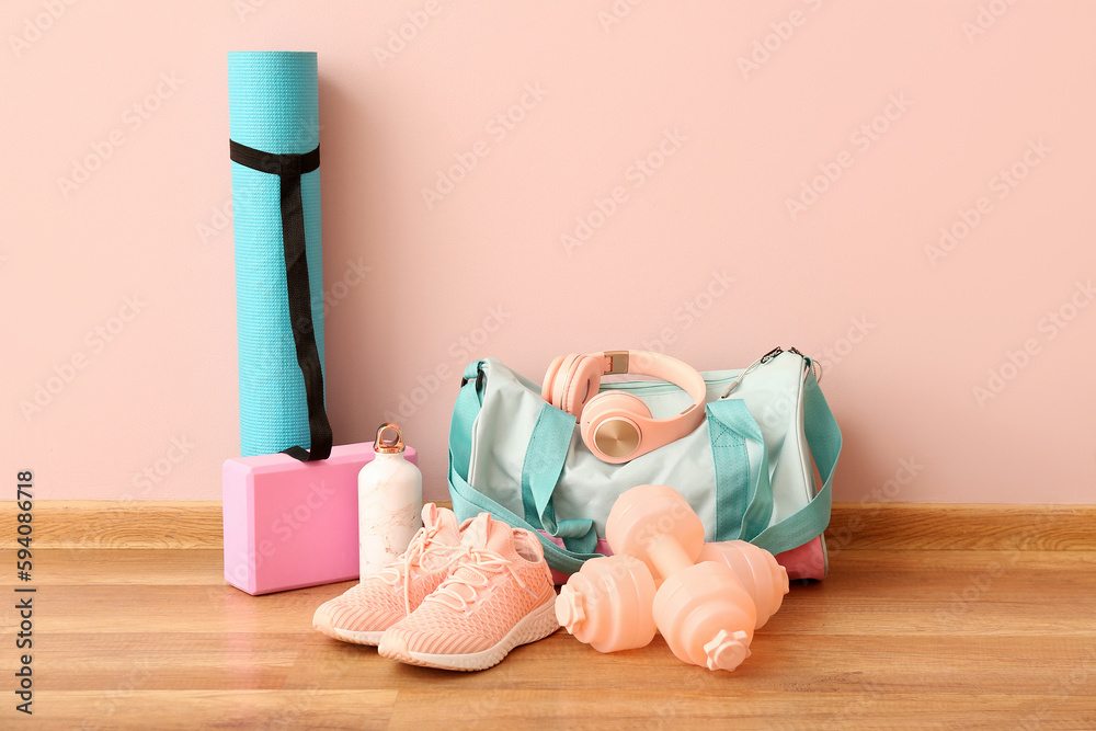 Sports bag with headphones, bottle, mat, sneakers and dumbbells near pink wall