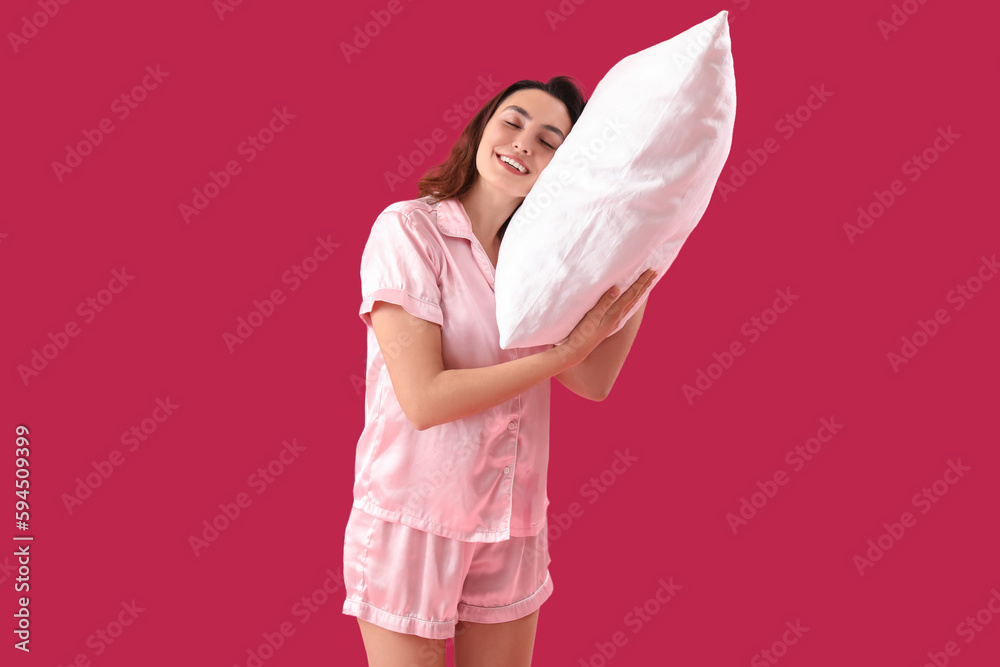 Young brunette woman in pajamas with pillow on pink background