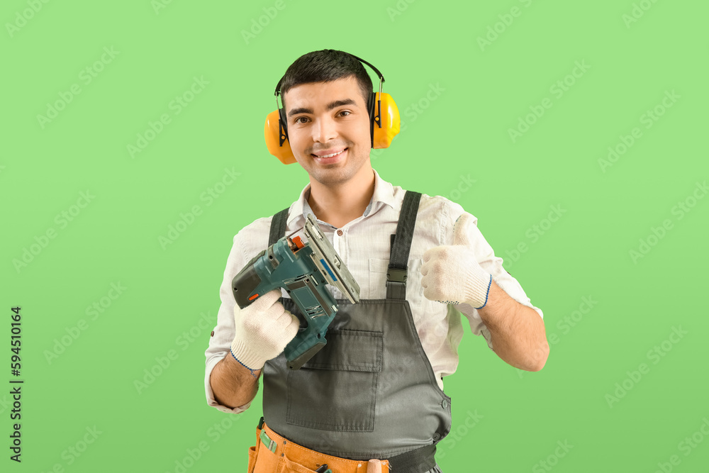 Young carpenter with cordless jigsaw showing thumb-up on green background