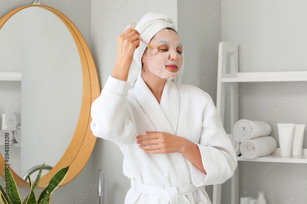 Young woman with sheet mask and facial massage tool in bathroom